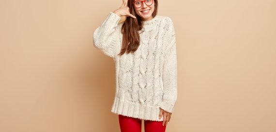 Call me back. Positive lovely woman does phone gesture, makes sign, asks being in touch, smiles happily, has friendly look, wears winter jumper, isolated over beige background. Body language concept