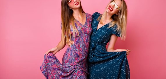 Couple of pretty best friends sister girls having fun together, wearing elegant summer pretty trendy dresses and sunglasses, posing at pink studio background, hugs and smiling, party atmosphere.